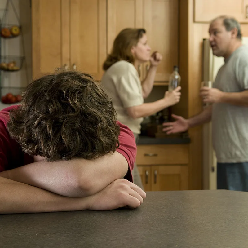 Picture of a child putting his head down on a table while his parents argue in the background. 