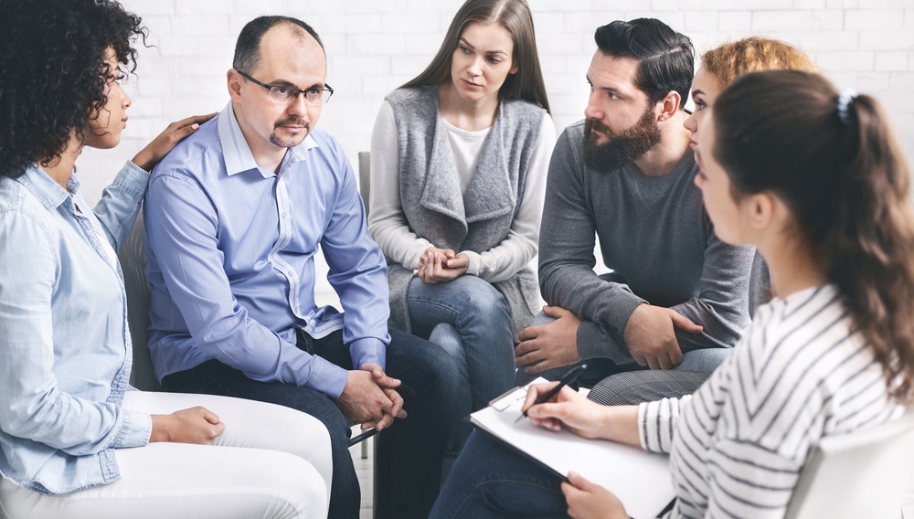 Concerned people comforting upset man at group therapy session meeting in rehab, panorama